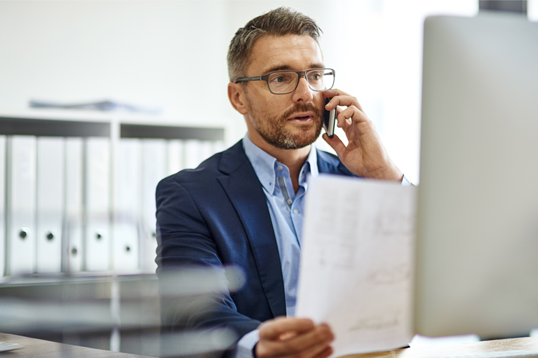 Businessman viewing critical data on both paper and desktop computer while talking on phone